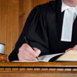 A judge sitting and writing on a paper which is placed on top of a wooden table, which is the header image of the blog entitled "choosing a criminal lawyer".