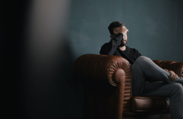 Man covering his head and eyes with his hand and is sitting on a brown couch, which is the header image of the blog entitled "professional association and misconduct inquiries"